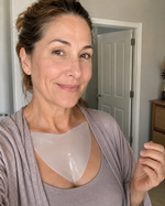 A woman wearing a translucent heart-shaped silicone or adhesive chest patch over a gray tank top and matching cardigan, standing indoors in front of a white door.